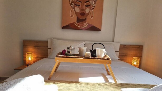 A neatly made bed with a tray of breakfast items, including a teapot, cups, and a book, in a room with a large painting on the wall and two bedside lamps. - Paramount Gardens Resorts D204 (Tersefanou)