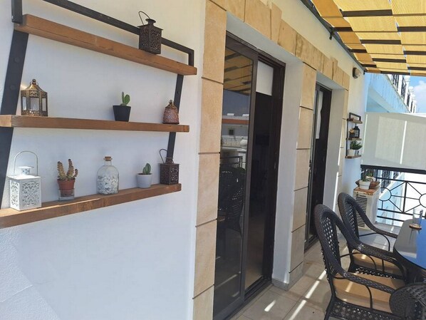 A balcony with wooden shelves holding decorative items, a glass door leading to an interior space, and a table with chairs. - Paramount Gardens Resorts D204 (Tersefanou)