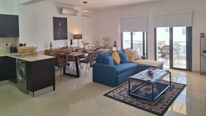 A spacious living area with a dining table, blue sofa, and a coffee table. There is a kitchen area with a washing machine and a flat-screen TV. - Paramount Gardens Resorts D204 (Tersefanou)