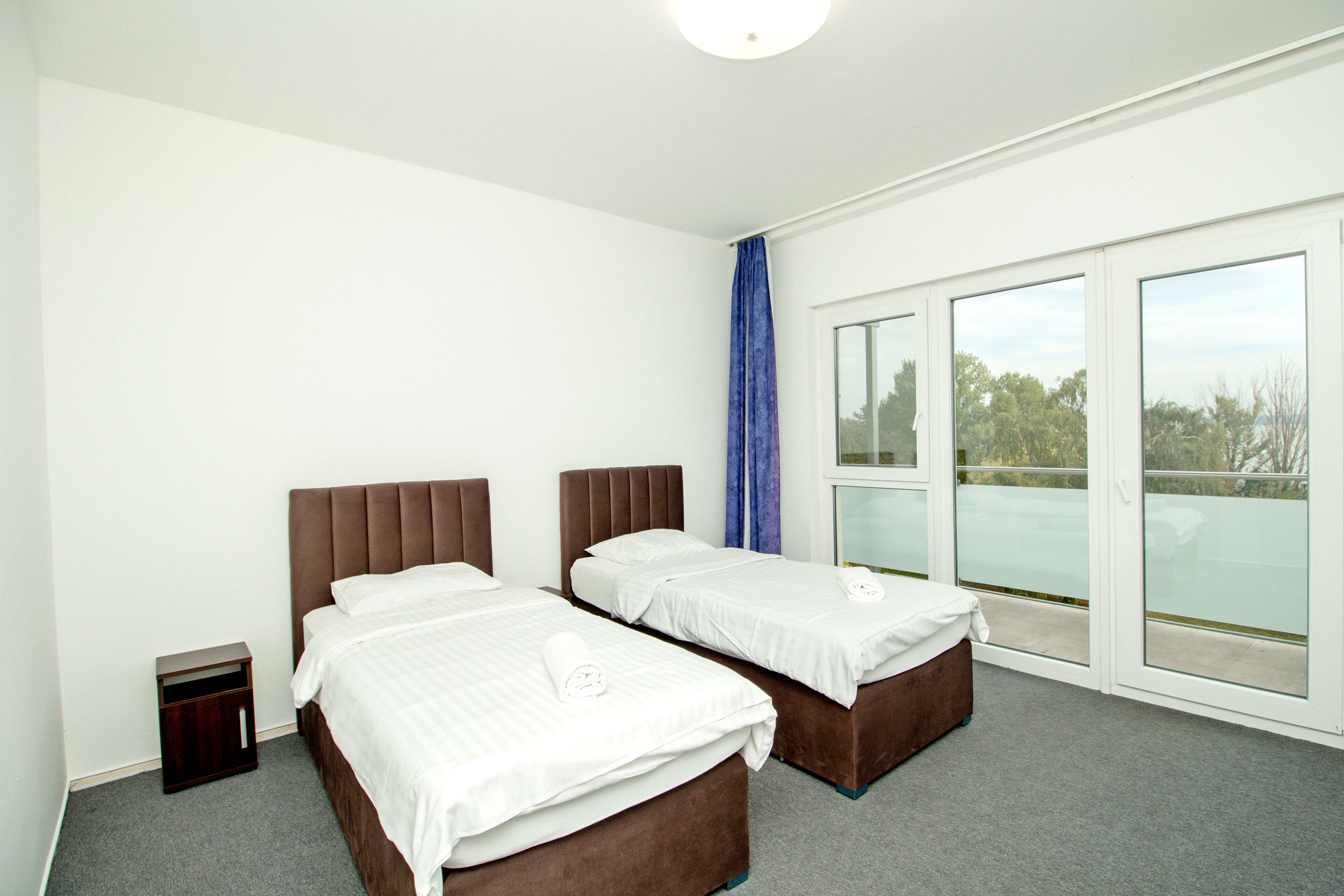 Quarto de hotel com duas camas, uma mesinha de cabeceira, uma janela grande com vista e uma luminária no teto.