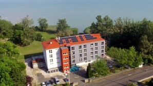 Un edificio de varios pisos con techo rojo y paneles solares, rodeado de follaje y una masa de agua al fondo.