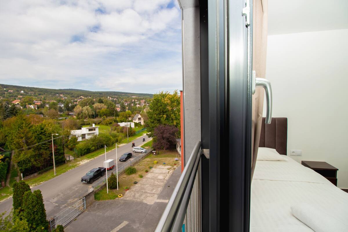 A hotel room with a bed, a window overlooking a street with cars and houses, and a view of a hilly landscape.