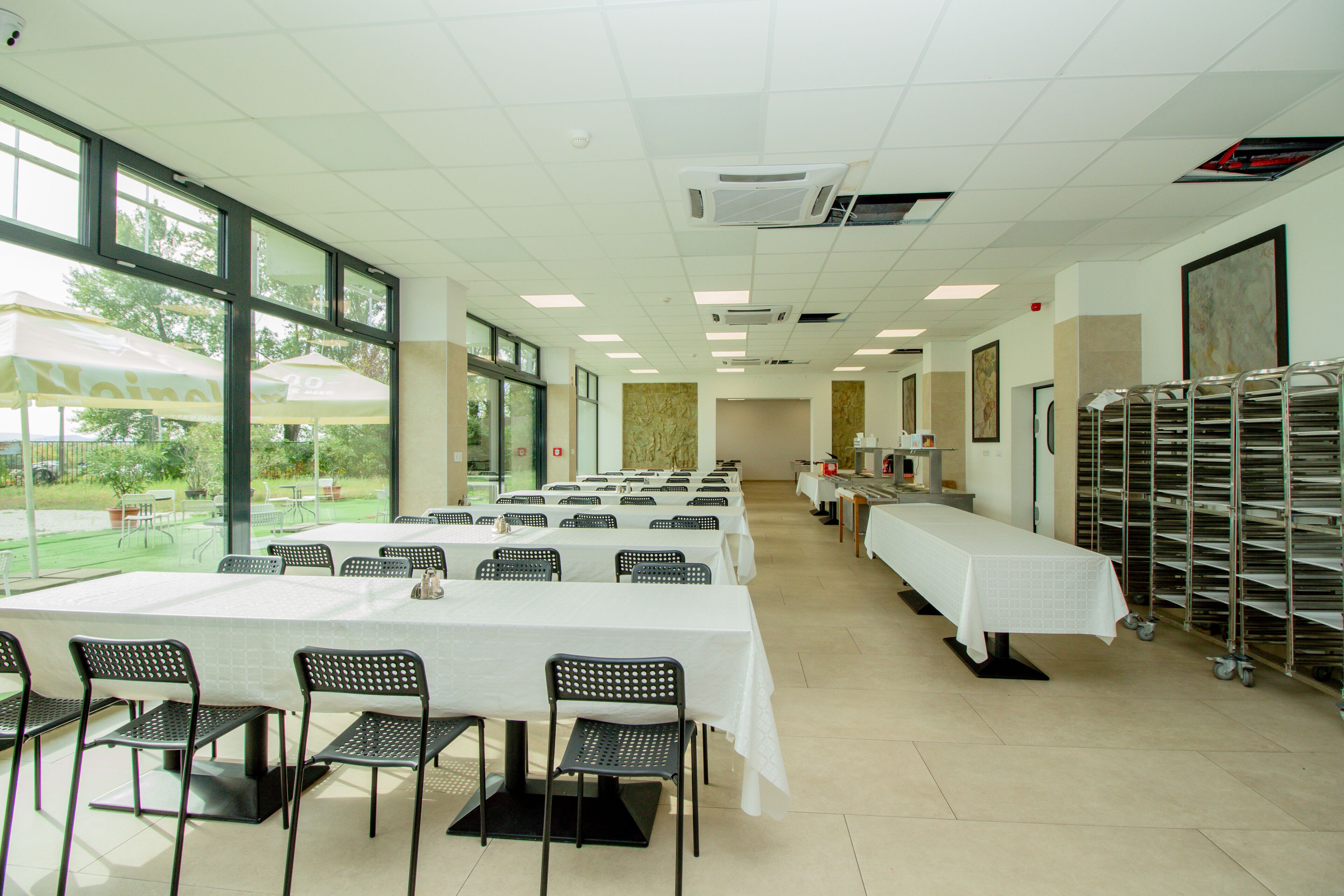 A spacious room with long tables and chairs, large windows, and a view of the outdoors.