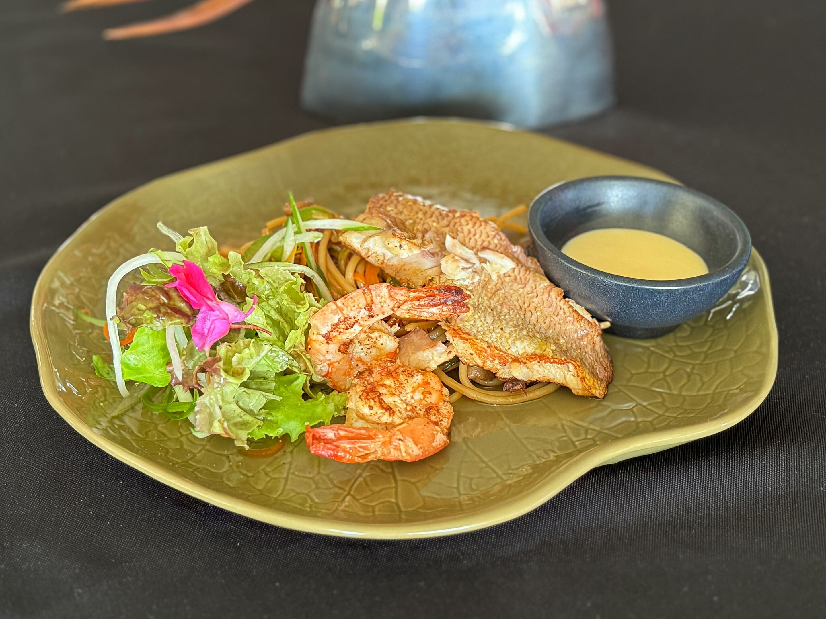 A plate of food with shrimp, fish, and salad, accompanied by a small bowl of sauce.