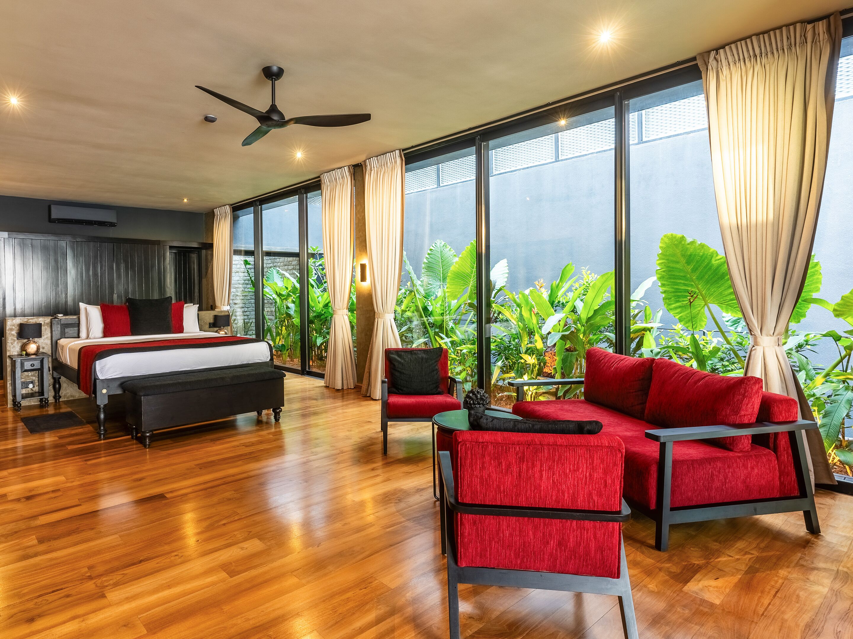 A modern hotel room with a large bed, a sofa, and a dining table. There are large windows with curtains, a ceiling fan, and a view of greenery outside.