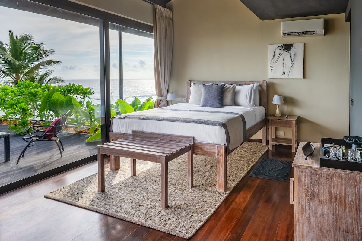 Une chambre avec un grand lit, une table de chevet en bois, une lampe de chevet, une vue sur l’océan et une fenêtre avec des rideaux.