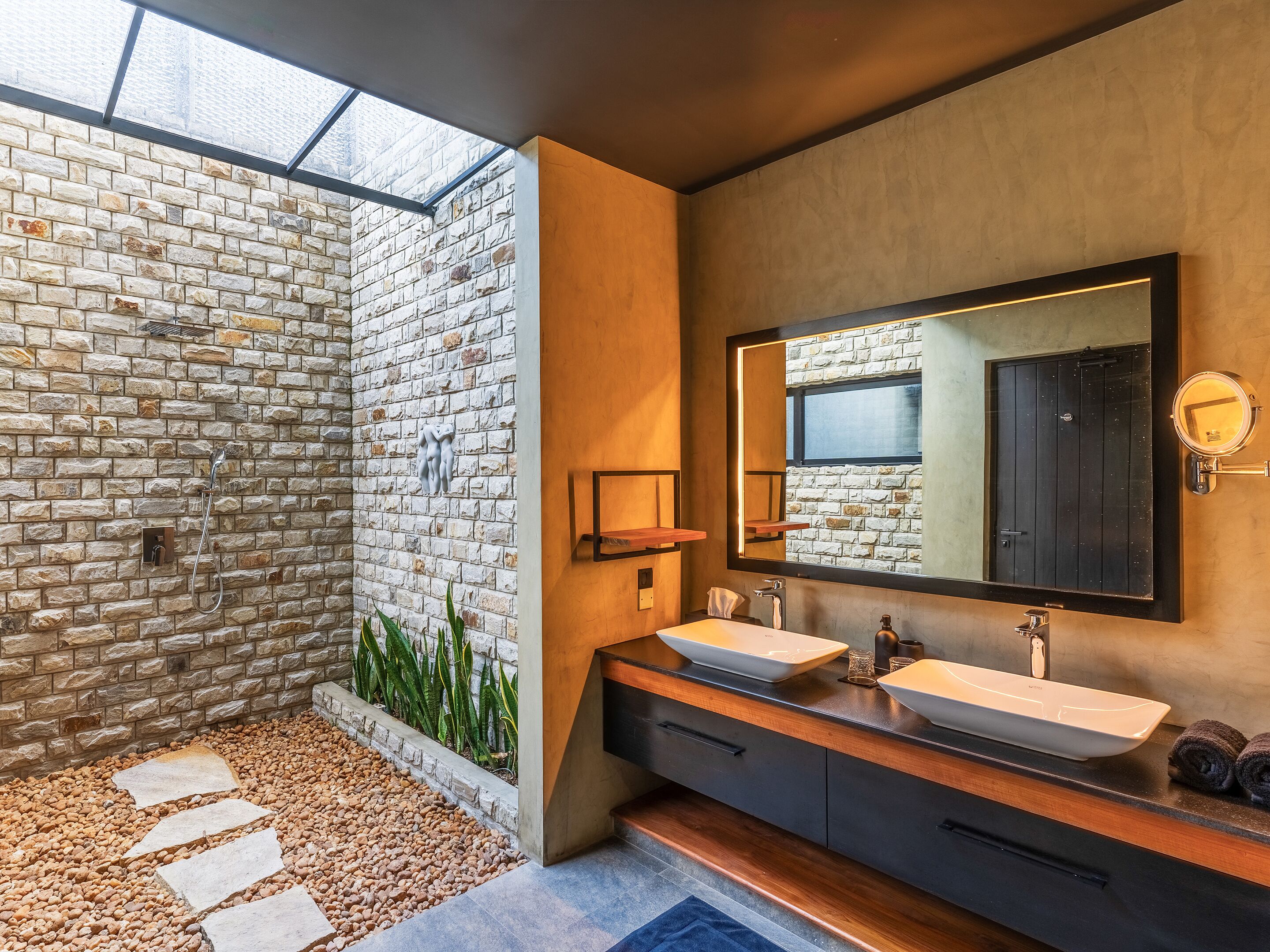 A modern bathroom with a stone wall, a skylight, a large mirror, and a dual sink vanity.