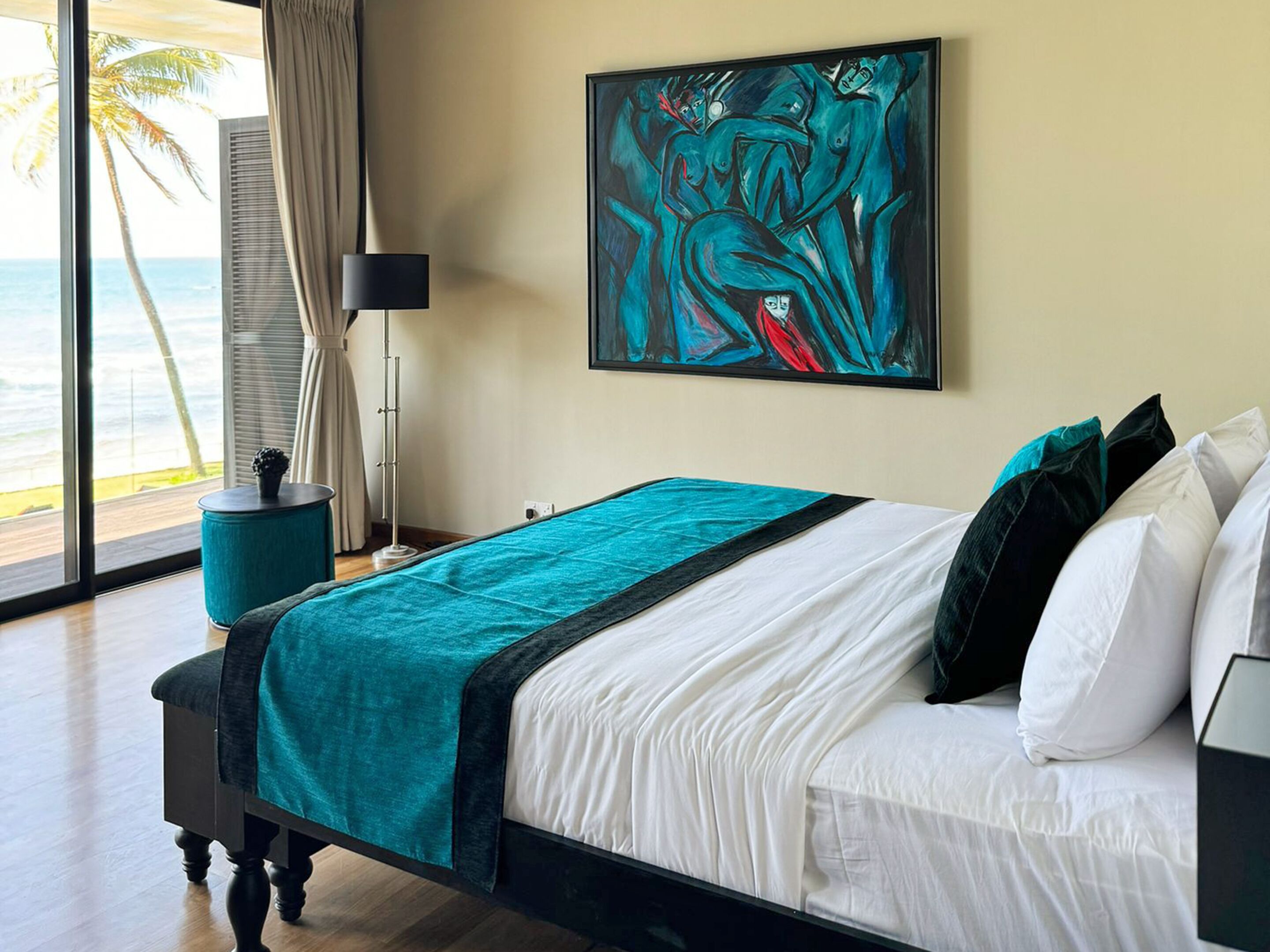 A hotel room with a bed, a painting, a lamp, and a view of the ocean.