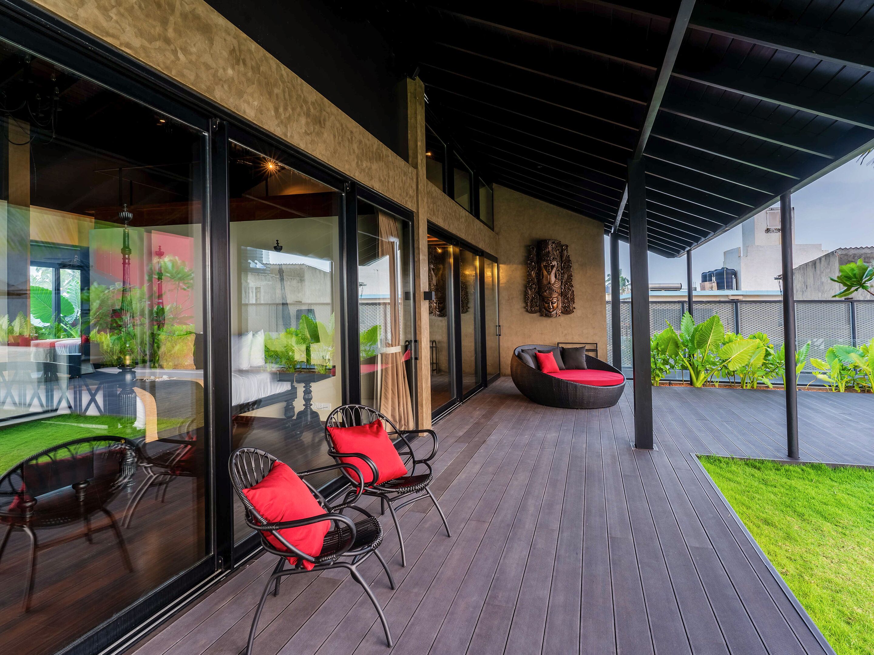 A modern outdoor seating area with wicker chairs and a cushioned bench, large glass doors leading to a lush garden, and a decorative wall piece.