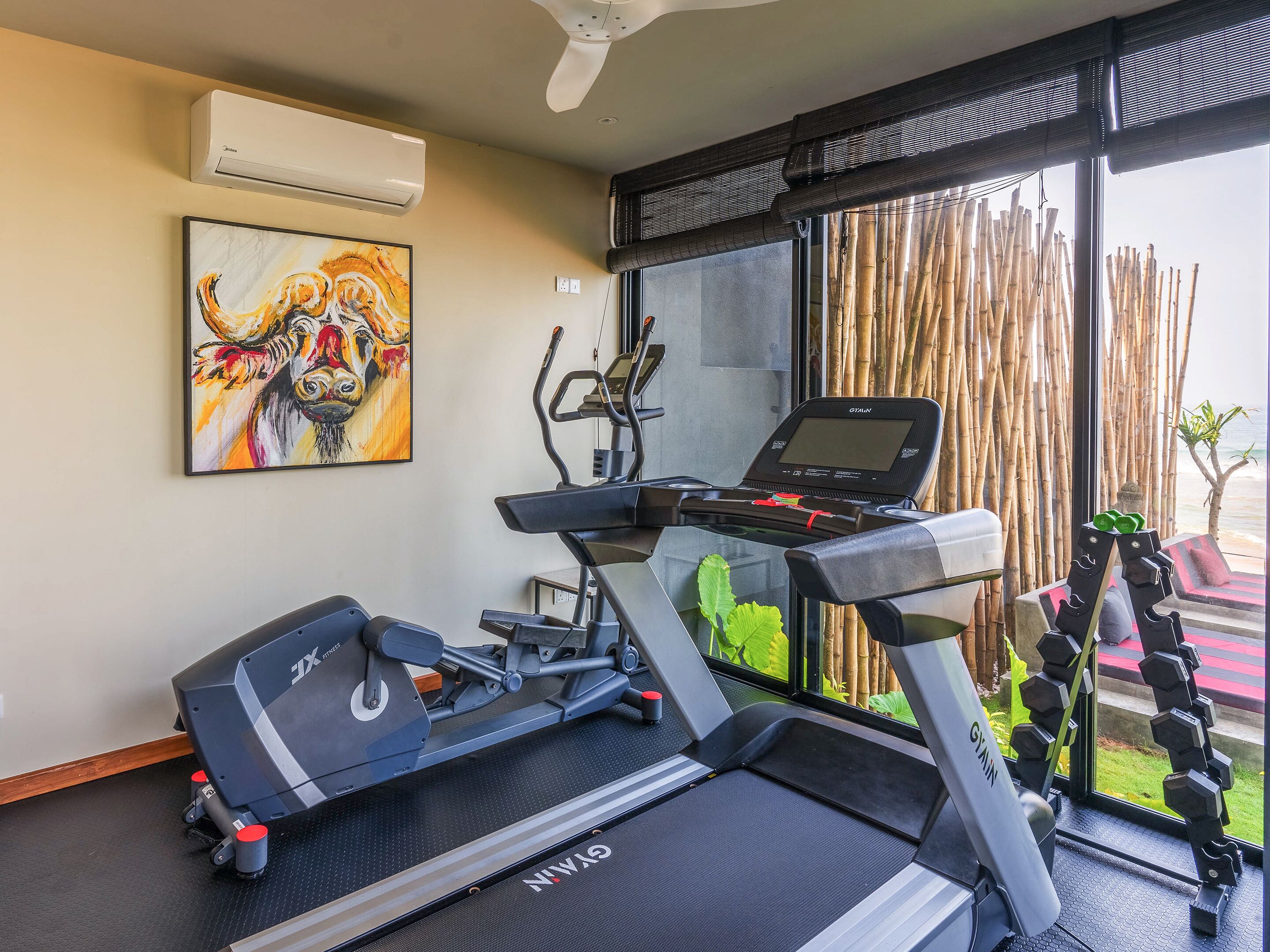 A gym with treadmills, an elliptical, and dumbbells. There is a painting on the wall and a window with bamboo blinds.