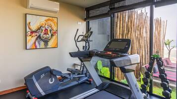 A gym with treadmills, an elliptical, and dumbbells. There is a painting on the wall and a window with bamboo blinds.