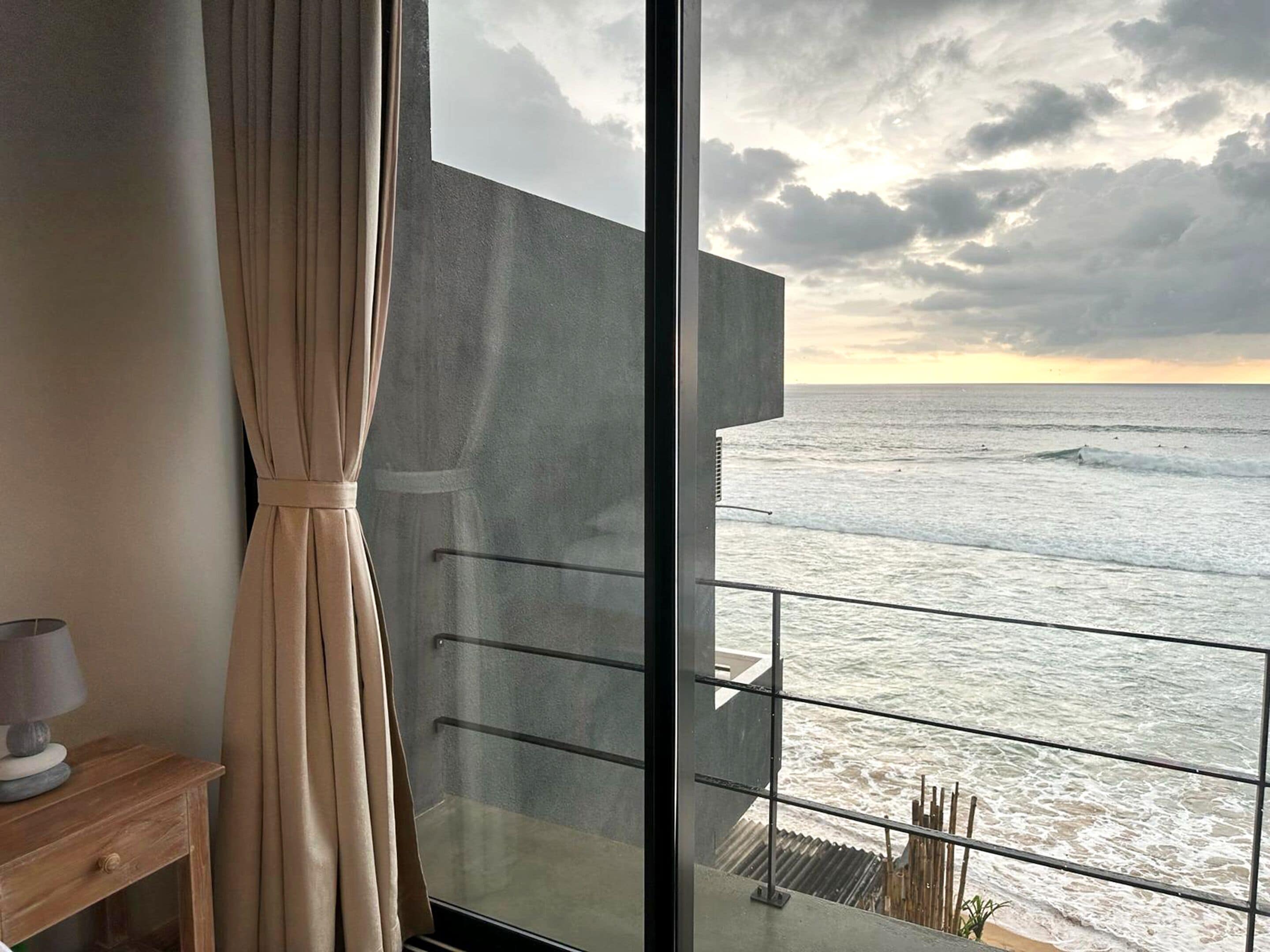 A room with a large window overlooking the ocean, a curtain, and a small wooden table with a lamp.