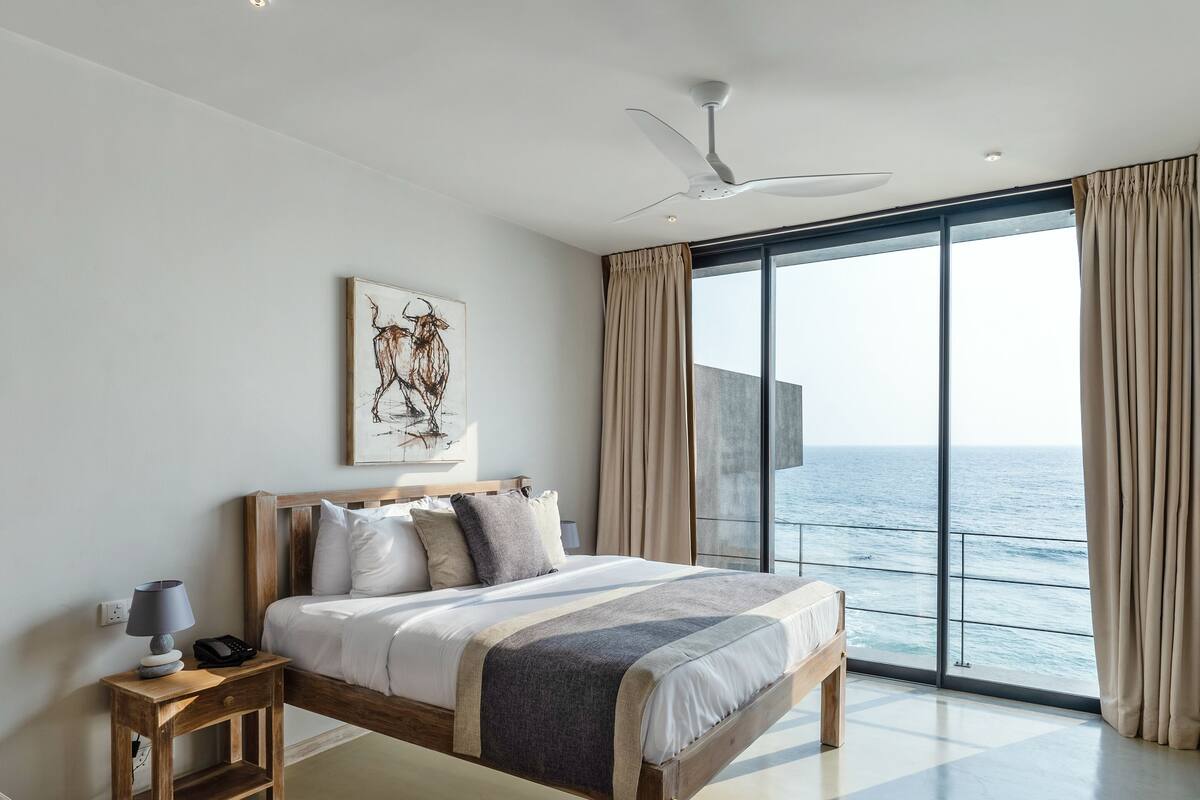 Une chambre à coucher moderne avec un grand lit, un ventilateur de plafond, une table de chevet avec une lampe et une vue sur l’océan grâce à de grandes fenêtres du sol au plafond.