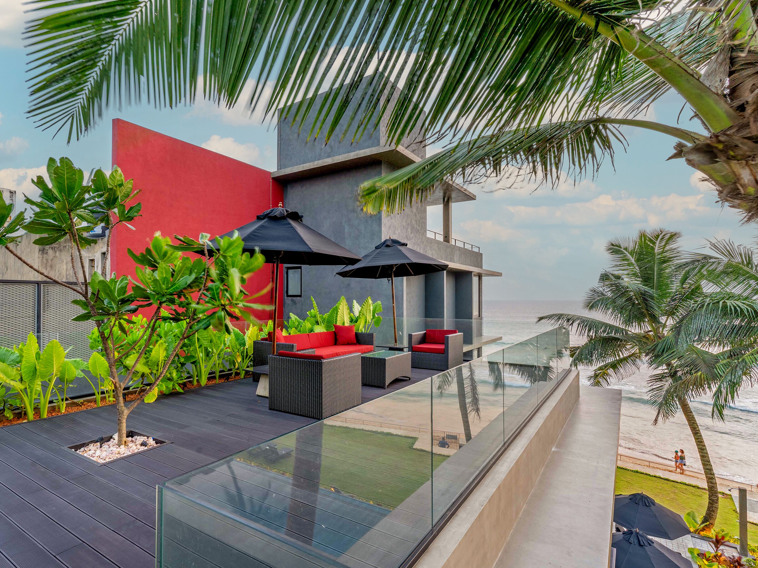 A modern outdoor seating area with glass railings overlooking the beach, featuring red cushions and black umbrellas.