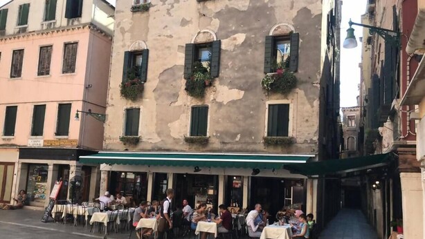 An outdoor cafe with tables and chairs, a waiter, and a narrow street between buildings. - Ca' Perdon Apartment (Venezia)