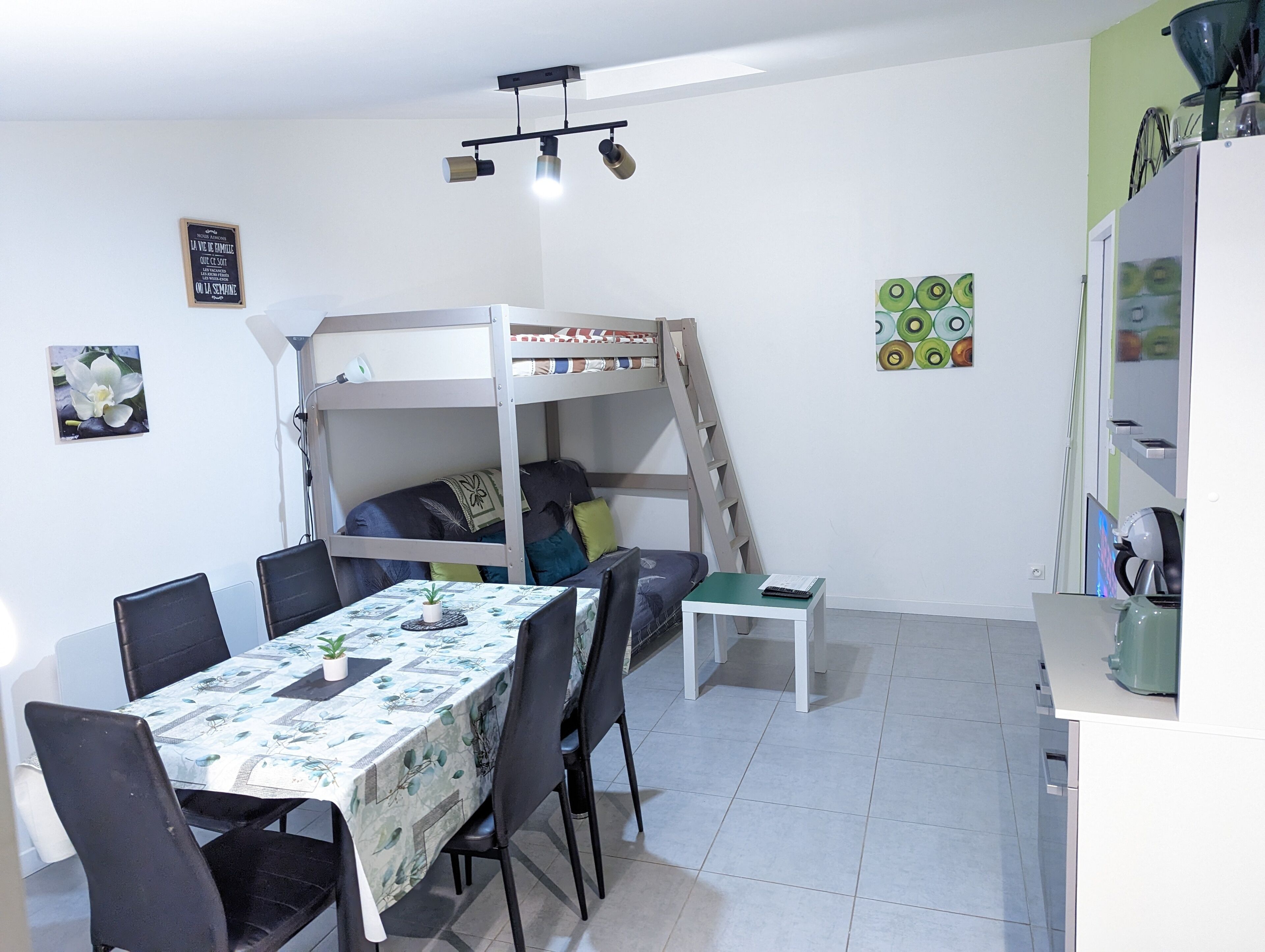 Habitación pequeña con literas, una mesa de comedor con sillas, una mesita blanca y una zona de cocina con refrigerador y lavamanos.