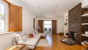 A spacious living room with a wooden floor, a white sofa with cushions, a stone fireplace, and a dining area in the background. - Casa Das Azeitonas (Azeitão)