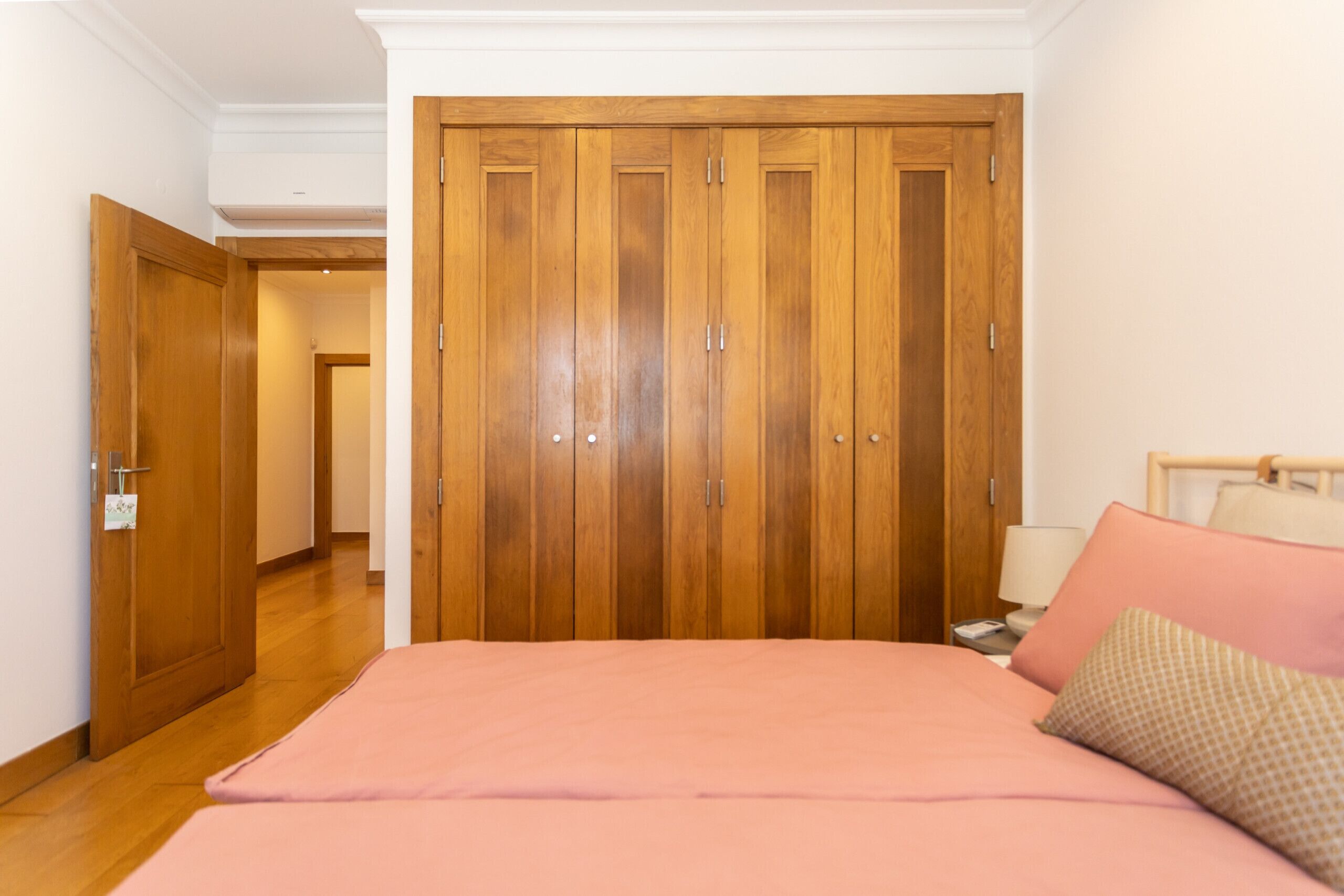 A bedroom with a wooden wardrobe, a bed with pink bedding, and a bedside table with a lamp.