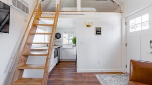 A loft bedroom with a wooden ladder, a bunk bed, a kitchen in the background, and a brown leather chair. - Urban Tiny House near Portland & Columbia Gorge (Camas)