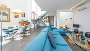 A modern living room with a blue sofa, white chairs, a glass coffee table, and a staircase leading to a balcony. - Casa Nilo Design - Villa with pool near the beach (Aroeira)