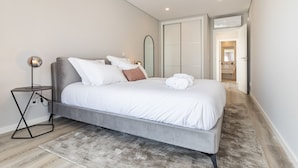 Une chambre moderne avec un lit gris, du linge de lit blanc, une petite table de chevet avec une lampe et un dressing.