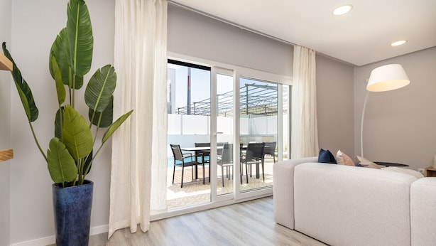 Un salon moderne avec un canapé, une plante en pot et une porte-fenêtre coulissante donnant sur une terrasse extérieure équipée d’une table et de chaises.