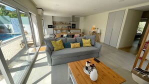 A modern living room with a grey sofa, wooden coffee table, and a view of the kitchen and pool area. - Casa Evae (Azeitão)
