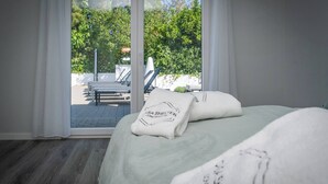 Ett sovrum med ett stort fönster, en säng med vita sängkläder och utsikt över en uteplats med solstolar.