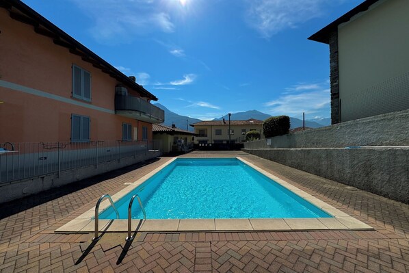 A swimming pool with a herringbone brick pattern surrounding it, adjacent to a building with a balcony and a stone wall.