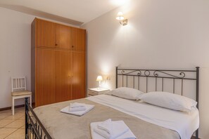 A neatly made bed with white linens, a wooden wardrobe, a bedside table with a lamp, and a chair.