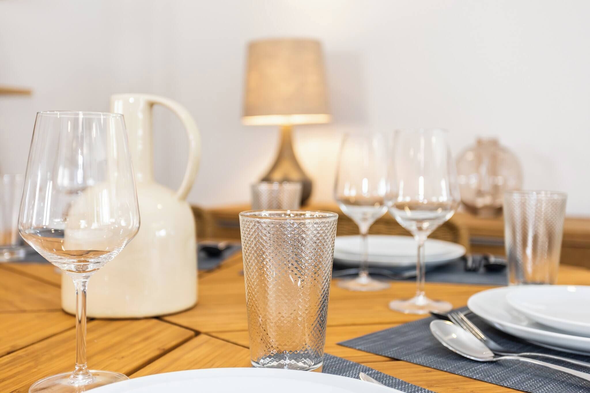 Une table à manger dressée avec de la vaisselle en verre transparent, un pichet et une lampe en arrière-plan.