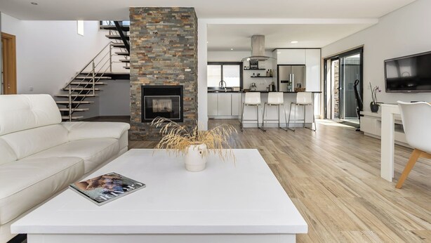 Un salon moderne avec un canapé blanc, une table basse blanche sur laquelle repose un magazine, une cheminée et, au fond, une cuisine.