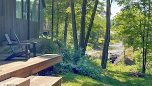 A wooden deck with a chair, overlooking a river and dense forest.
