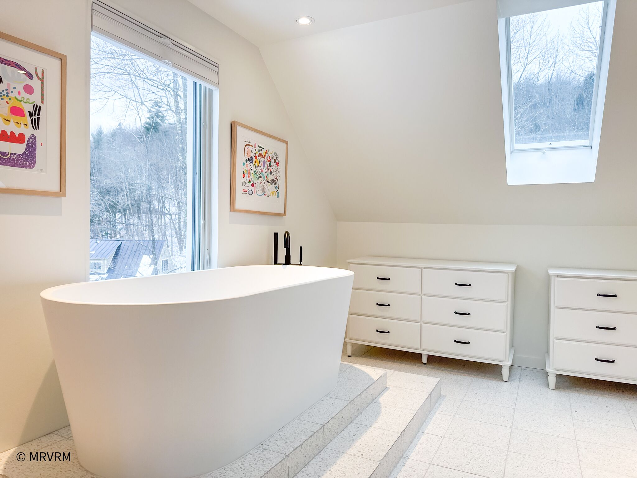 A modern bathroom with a freestanding bathtub, white cabinetry, and large windows providing natural light.