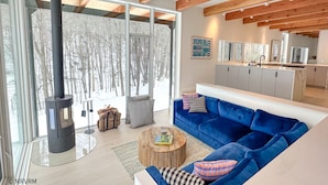 A modern living room with a blue sofa, a round wooden coffee table, and a large window overlooking a snowy forest.