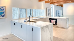 A modern kitchen with a marble island, wooden beams, and large windows.