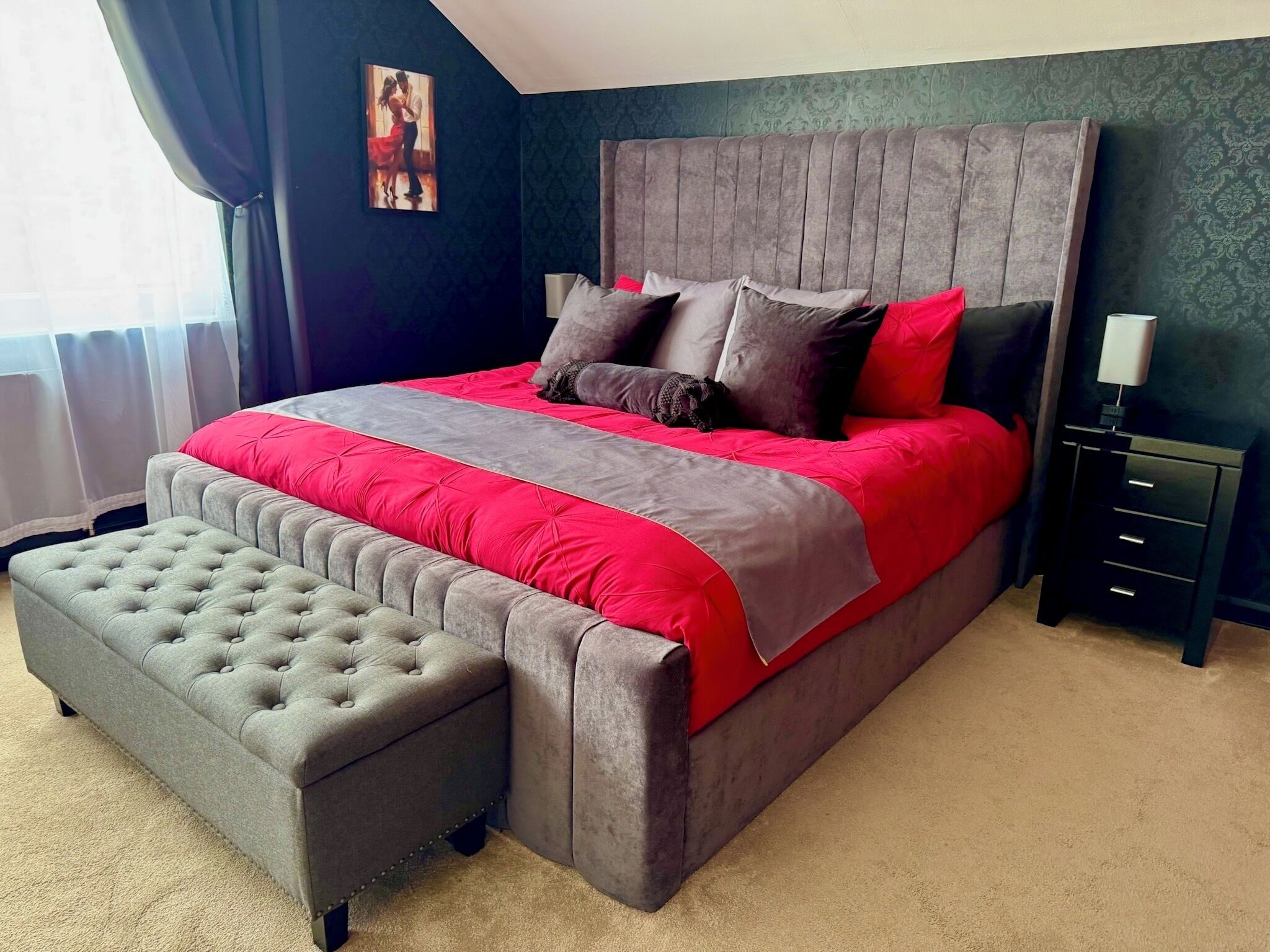 A bedroom with a large bed, grey upholstered bench, dark walls, and a framed picture on the wall.