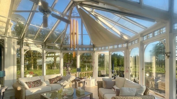 Une véranda meublée de mobilier en osier, dotée de tables en verre et offrant une vue sur l’extérieur.