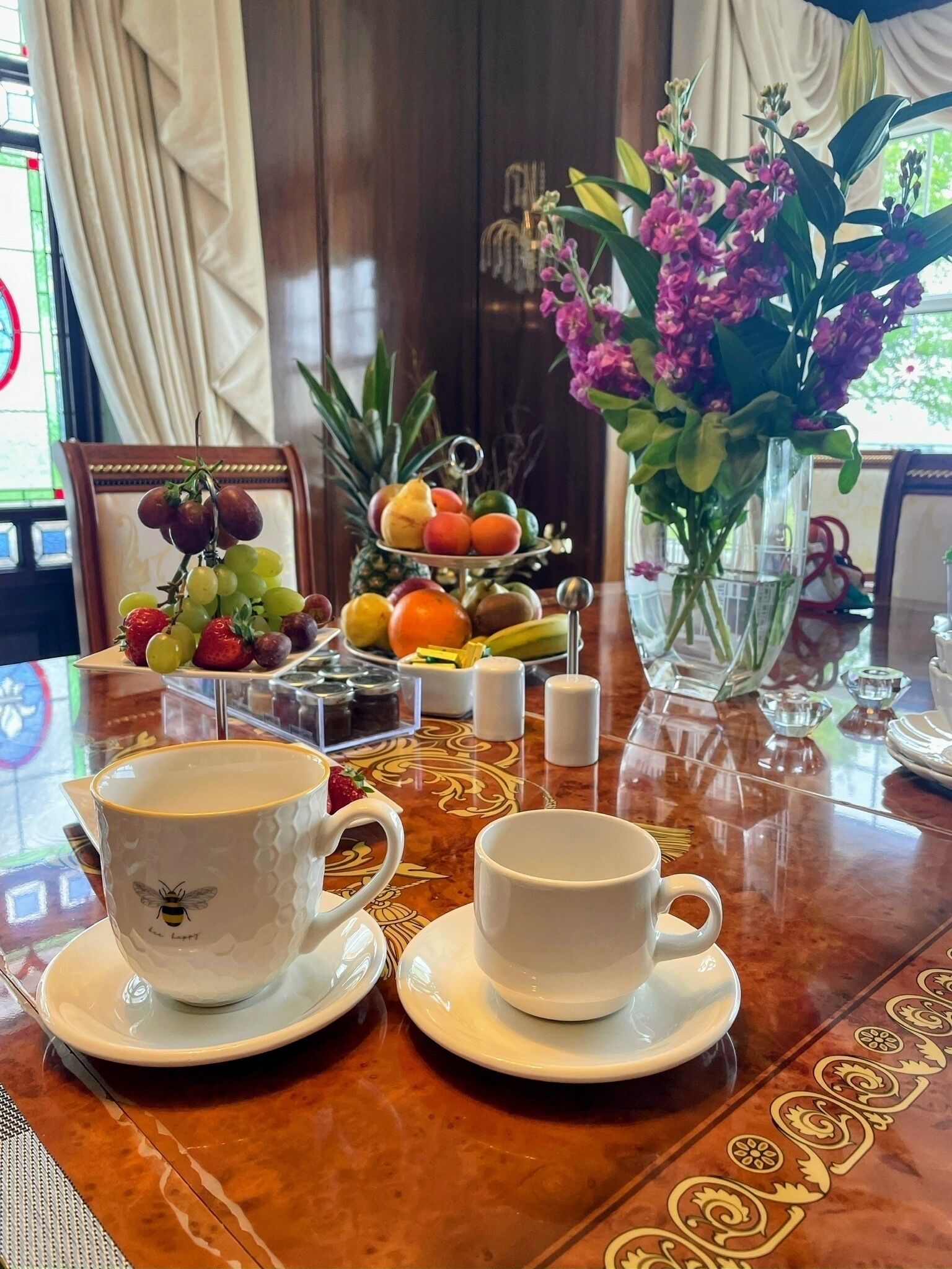 Ein Tisch mit zwei weißen Tassen und Untertellern, einer Obstplatte und einer Vase mit violetten Blumen.