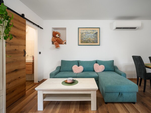 A teal sofa with heart-shaped cushions, a white coffee table, a teddy bear in a niche, a framed landscape painting, and a modern air conditioning unit.