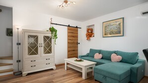 Un soggiorno moderno con pavimento in legno, un mobile bianco con ante in vetro, un divano color petrolio con cuscini a forma di cuore, un tavolino e una lampada a sospensione.