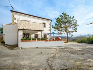 Une maison à deux étages avec une façade en pierre, un balcon orné de plantes en pot et une voiture rouge garée dans l’allée.