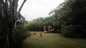 Une petite cabane située dans une zone boisée, avec une personne debout dans la cour.