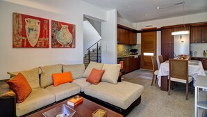 A living room with a sofa, coffee table, and artwork on the wall. There is a staircase leading to a kitchen area with wooden cabinets and a dining table. - Bali private pool villa overlooking the lovely bay (Vlichada)