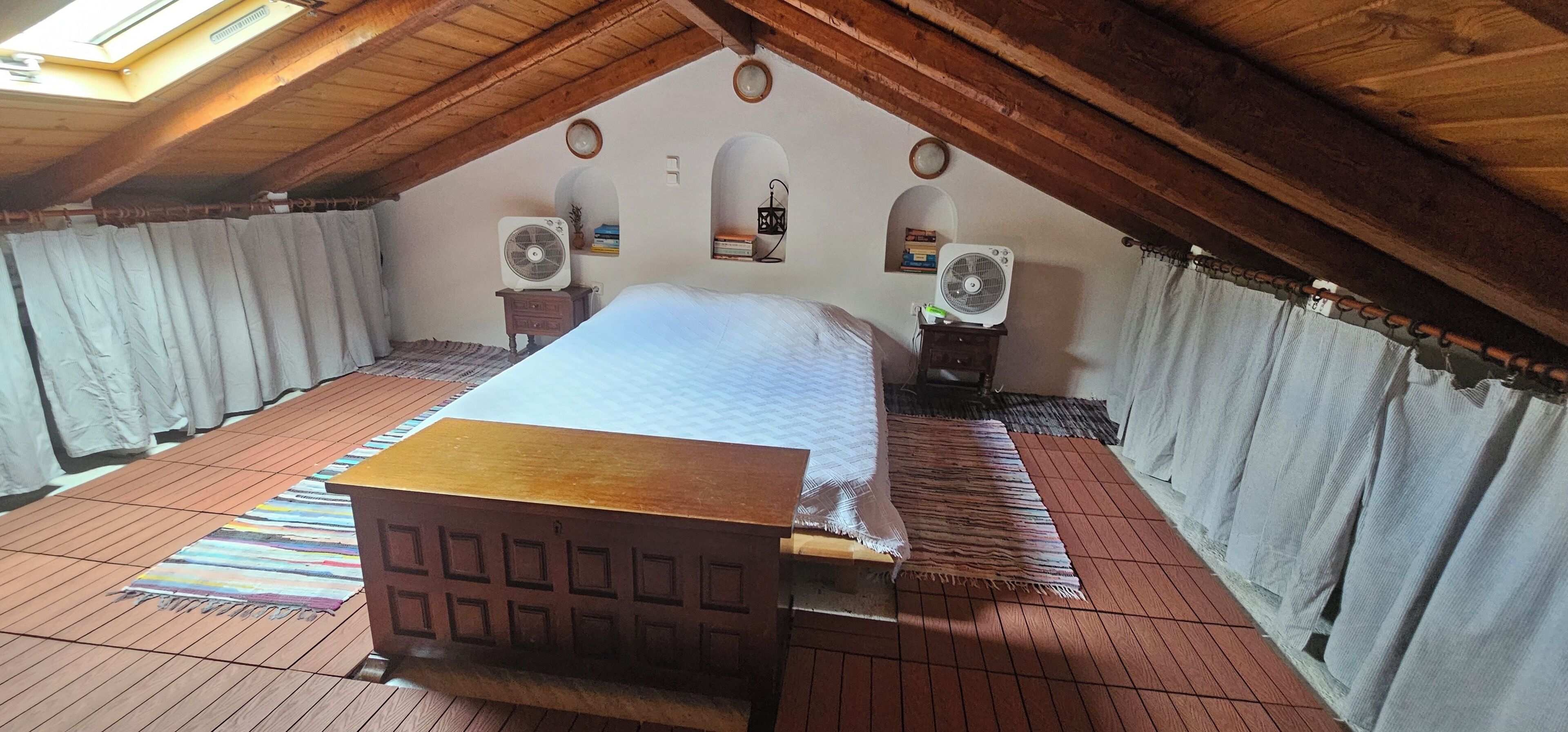 Una camera da letto con soffitto in legno, un letto, due comodini, due ventilatori e un lucernario.