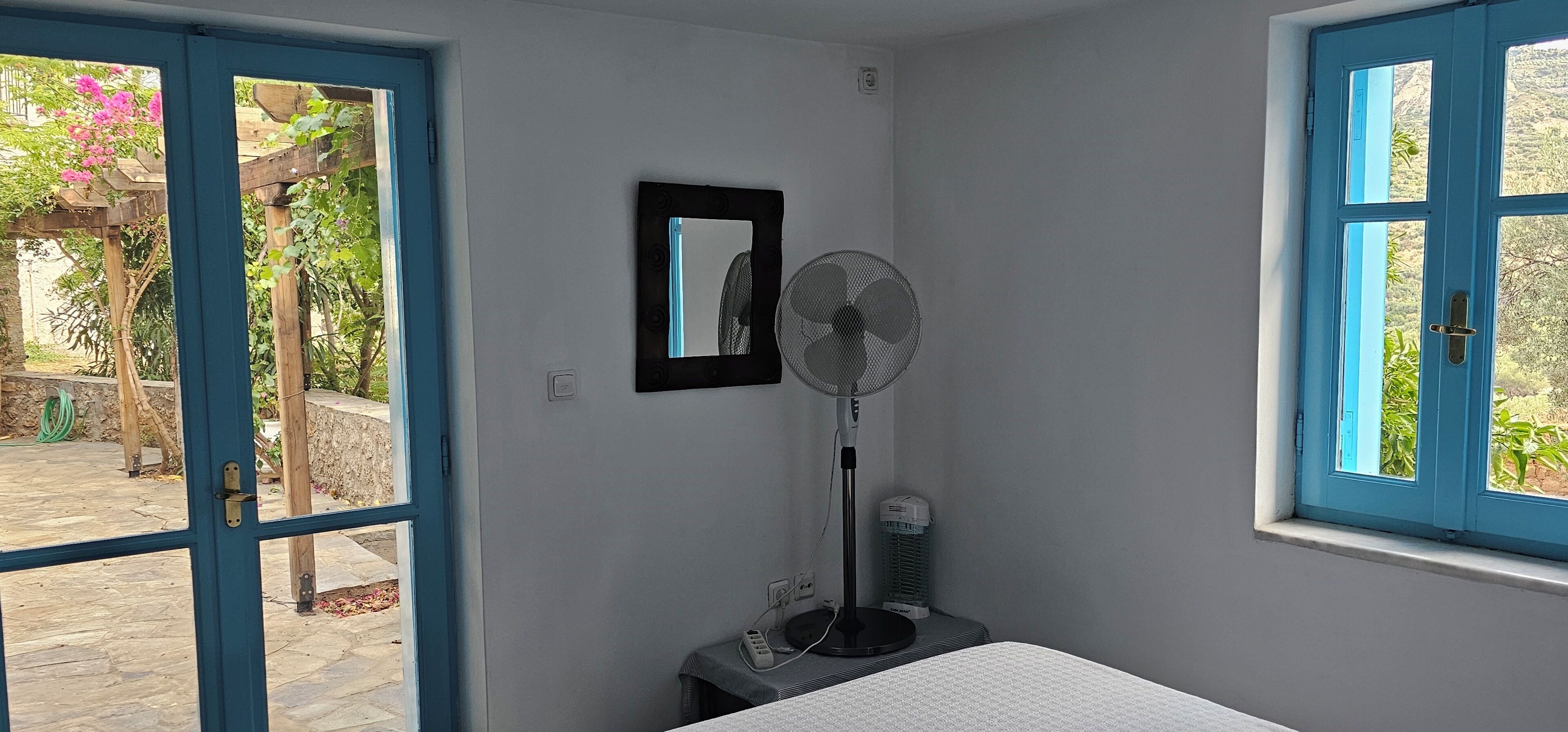 Una habitación con una cama, un ventilador, un espejo y una ventana con vistas a la vegetación.