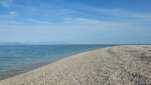 Una spiaggia di ciottoli con acqua calma e montagne in lontananza sotto un cielo sereno.