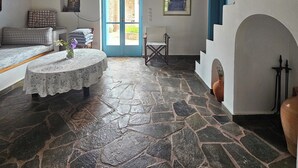 Una habitación con suelo de piedra, techo de madera, una puerta azul, pared blanca, cama, una mesa con mantel de encaje y una silla.