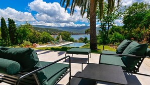 Un espace extérieur aménagé avec des fauteuils confortables et une table, offrant une vue sur une piscine et un paysage pittoresque composé d’un lac et de montagnes.