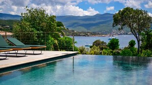 Une piscine d’eau cristalline, bordée de chaises longues, avec vue sur la mer et les montagnes.